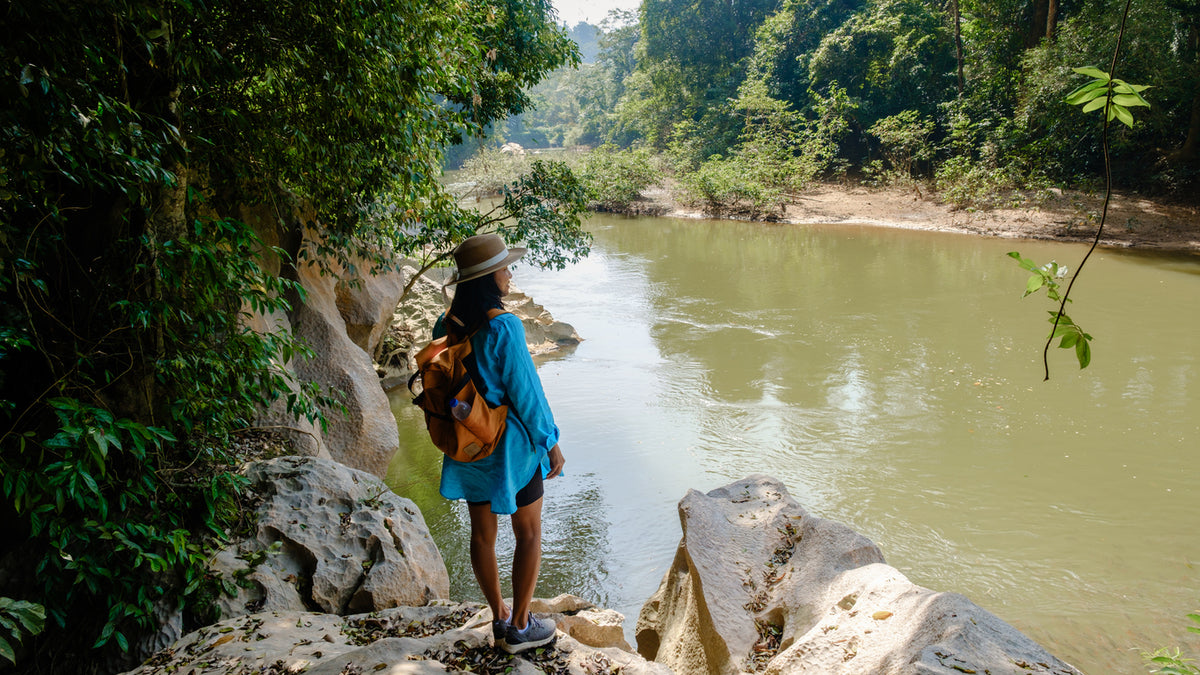 A woman stands on a rocky riverside path wearing a blue dress, brown hat, and orange backpack, looking out at a wide river surrounded by dense green forest.