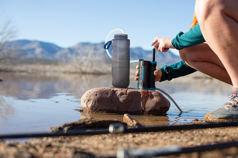 A person crouches beside a body of water outdoors, using a LifeSaver Wayfarer connected to a gray bottle with tubing. A large rock sits in the water, and mountains are visible in the background.